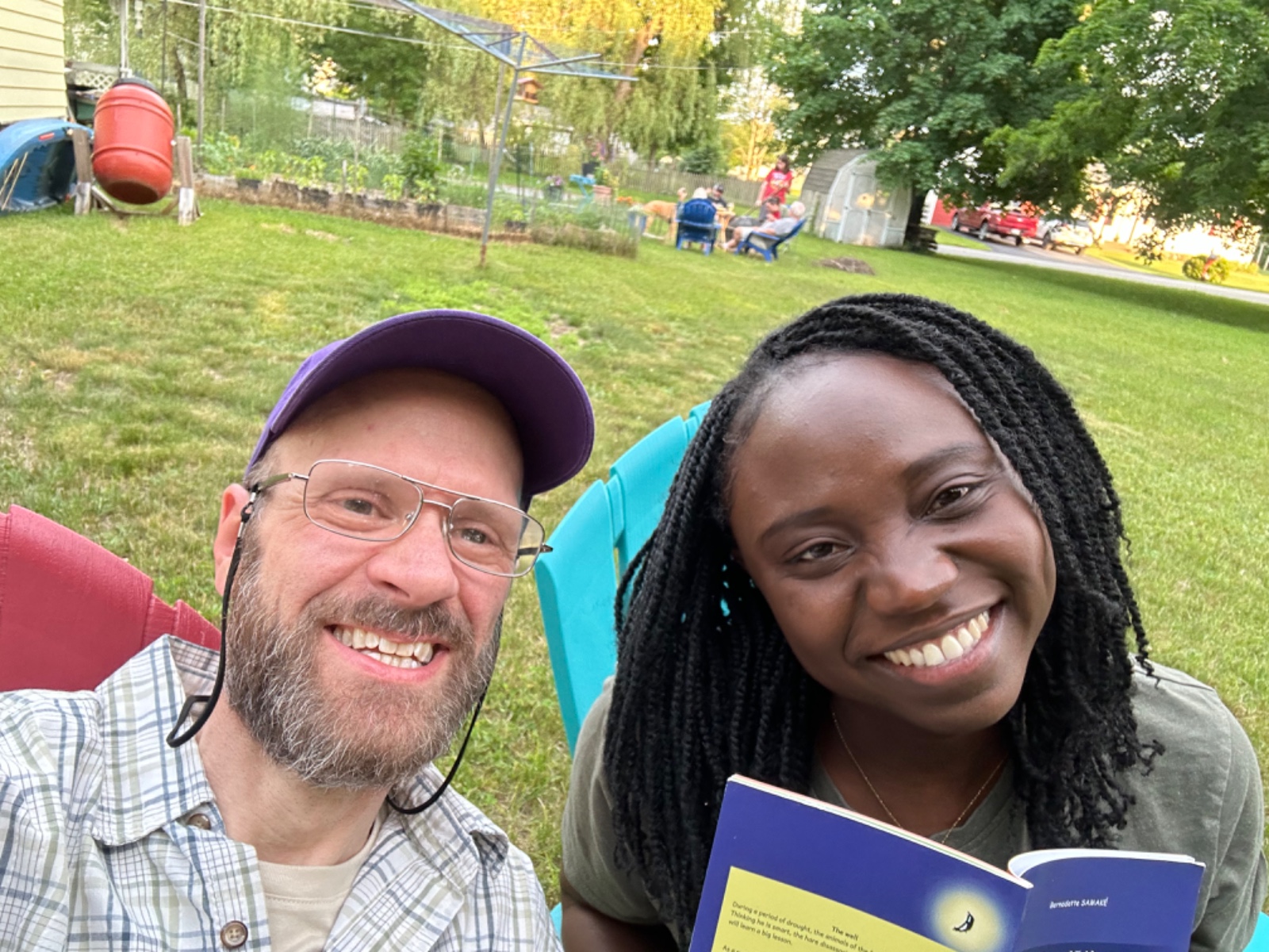 Michael Bleau wearing reading glasses and seated outdoors beside an author holding her published book.