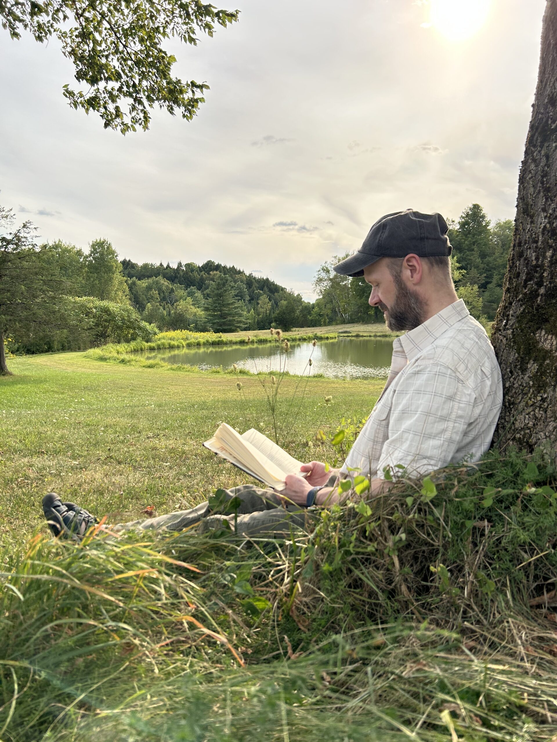 Michael Bleau sitting outdoors and reading a book while leaning against a tree near a pond.