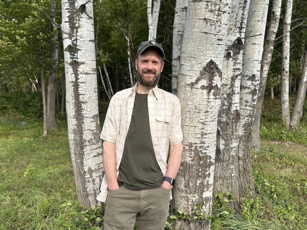 Close-up portrait of Michael Bleau leaning against a birch tree outdoors in a wooded setting.