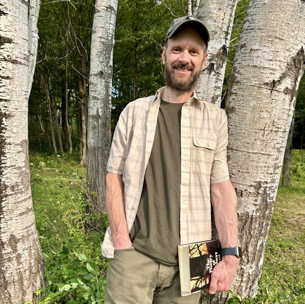 Michael Bleau standing outdoors among birch trees, holding a book and smiling.