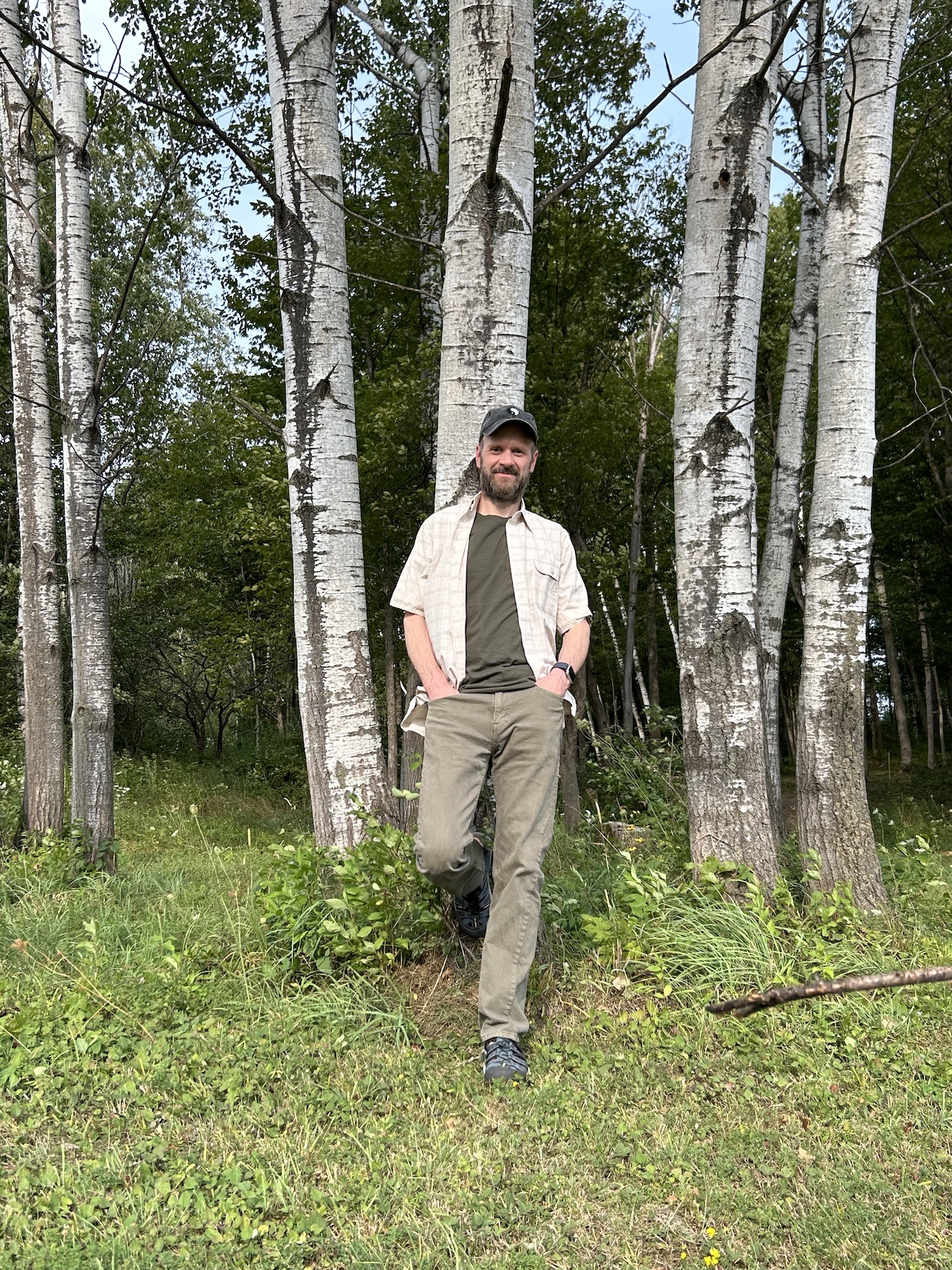 Michael Bleau standing outdoors with one foot against a birch tree, wearing casual clothing in a wooded setting.